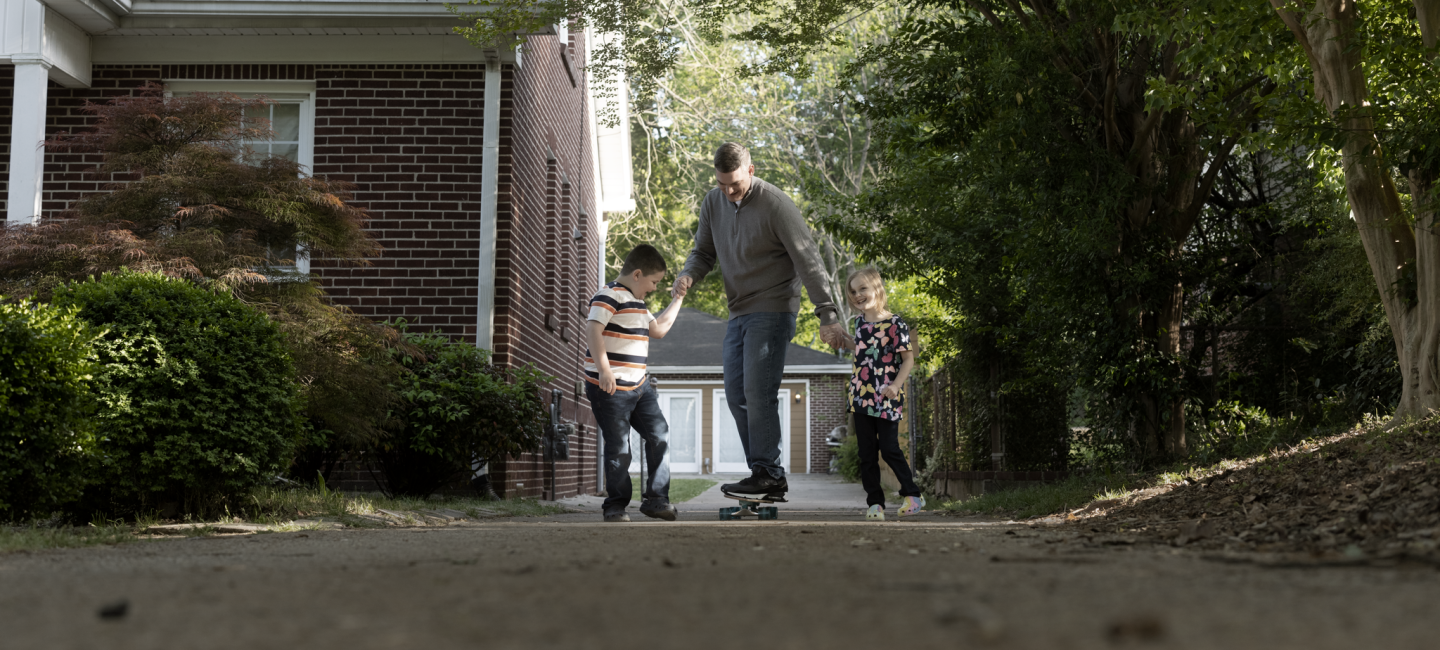 Father outside on a skateboard holding his two kids hands